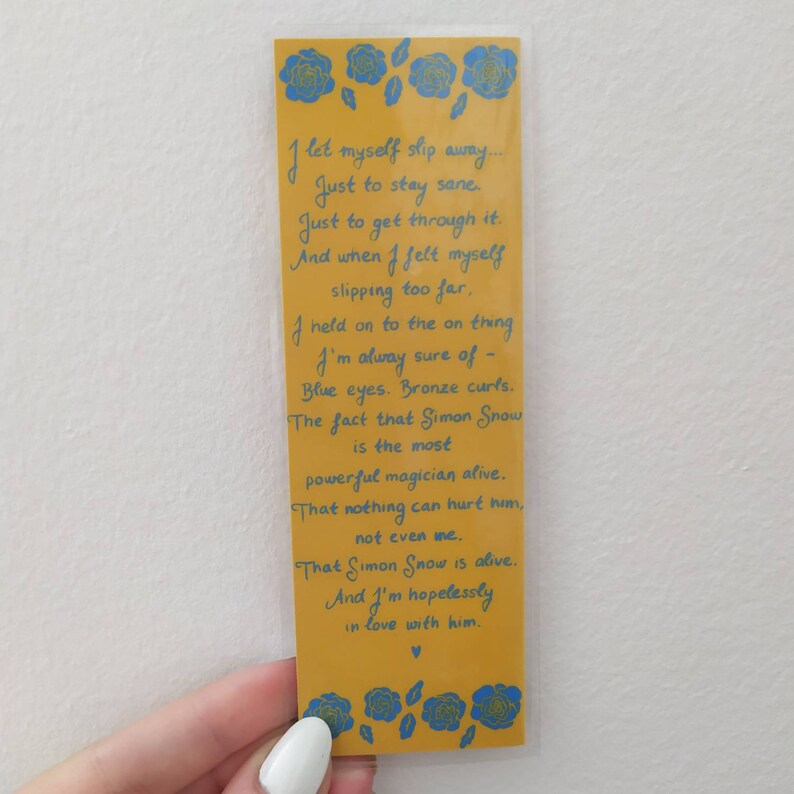 May include: Yellow bookmark with blue floral designs and a handwritten poem about a fictional character named Simon Snow. The poem reads: "I let myself slip away... Just to stay sane. Just to get through it. And when I felt myself slipping too far. I held on to the one thing I'm always sure of - Blue eyes. Bronze curls. The fact that Simon Snow is the most powerful magician alive. That nothing can hurt him, not even me. That Simon Snow is alive. And I'm hopelessly in love with him."