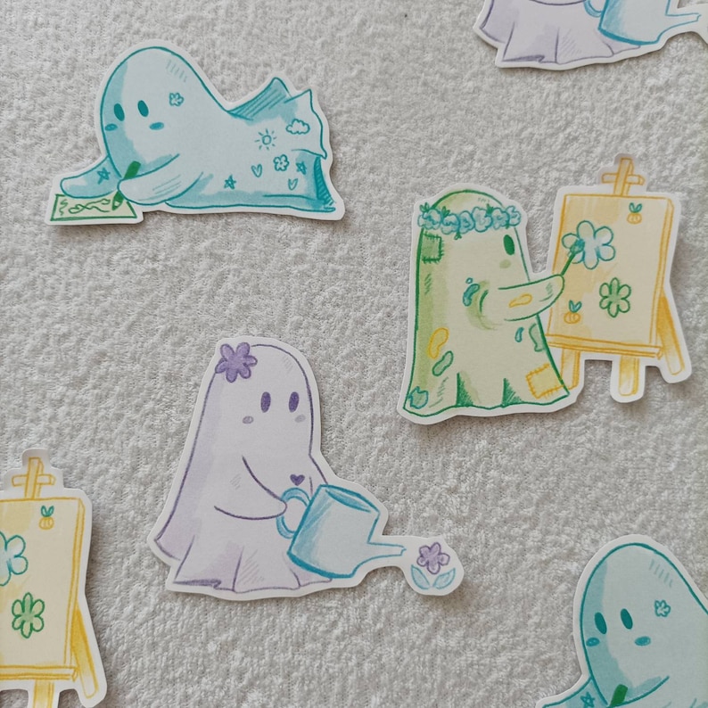 May include: Four cute ghost stickers with different designs. One ghost is holding a paintbrush and standing next to an easel with a flower painting. Another ghost is watering a flower with a watering can. One ghost is writing on a piece of paper and another ghost is wearing a flower crown.