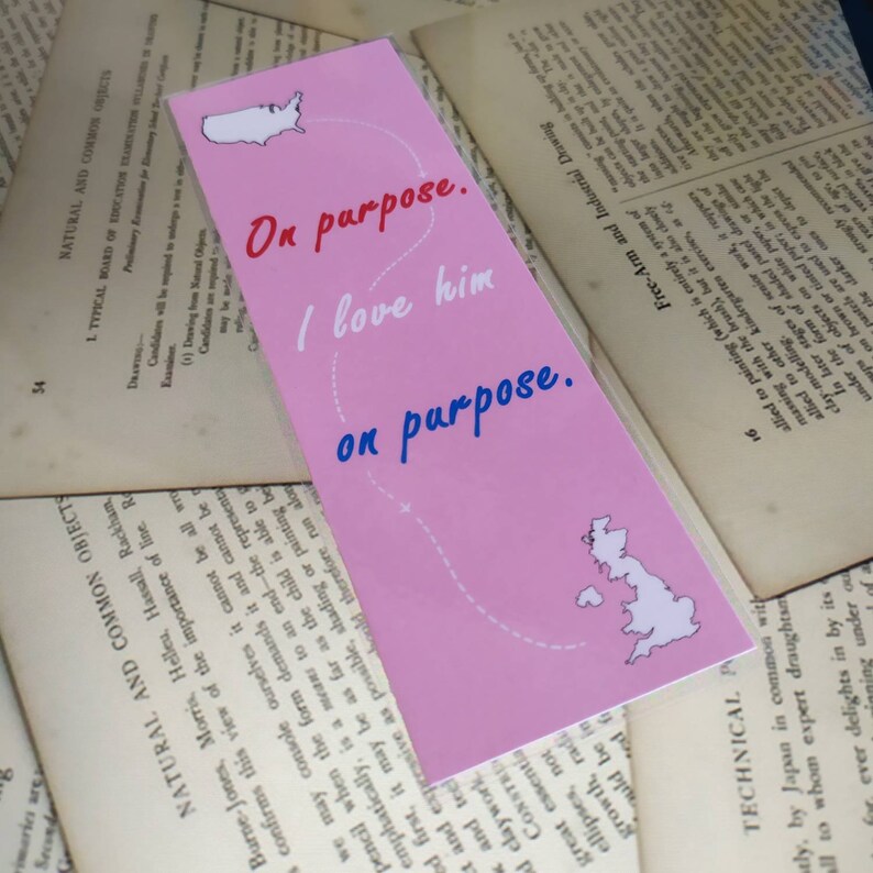May include: A pink bookmark with a white outline of the United States and the United Kingdom. The bookmark has the text "On purpose. I love him on purpose." in blue and white text.