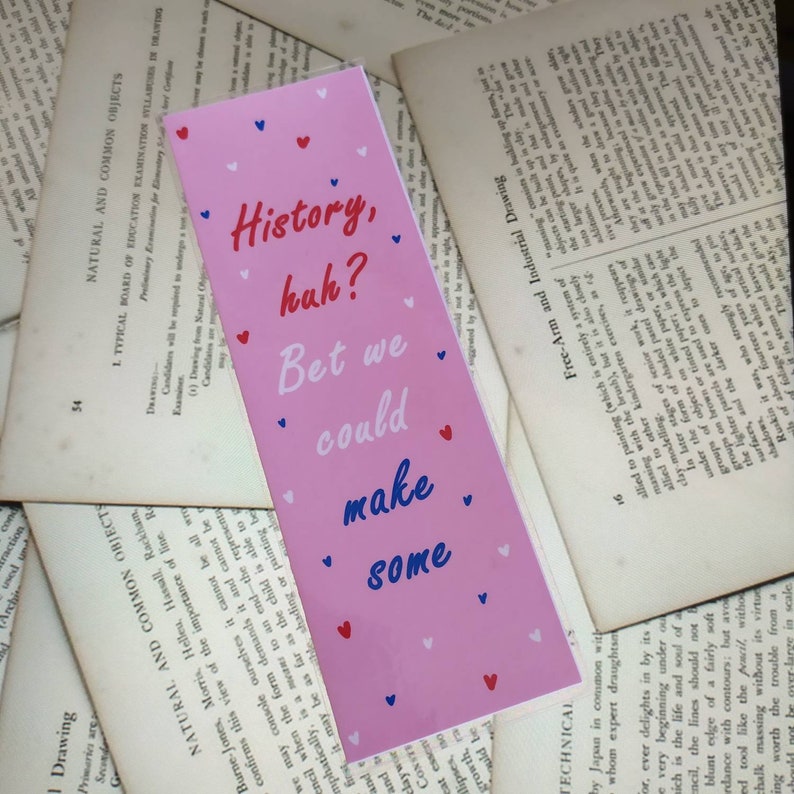 May include: Pink bookmark with the text "History, huh? Bet we could make some" in blue and white lettering. The bookmark is decorated with small red and white hearts.