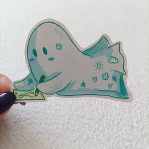 May include: A blue ghost sticker with a green pen, drawing on a piece of paper. The ghost has a cloud, sun, stars, and hearts drawn on it.