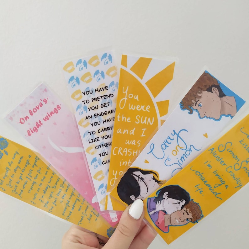 May include: A set of six bookmarks featuring colorful illustrations and quotes. The bookmarks are printed on white cardstock and have a glossy finish. The quotes include: "On love's light wings", "You have to pretend you get an endgame", "You were the sun and I was crashing into you", "Carry on Simon", "Simon Snow", and "Aleister Crowley I'm living a charmed life."