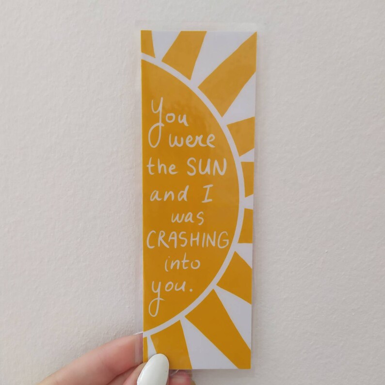 May include: A yellow bookmark with a white sun design and the text "You were the sun and I was crashing into you."
