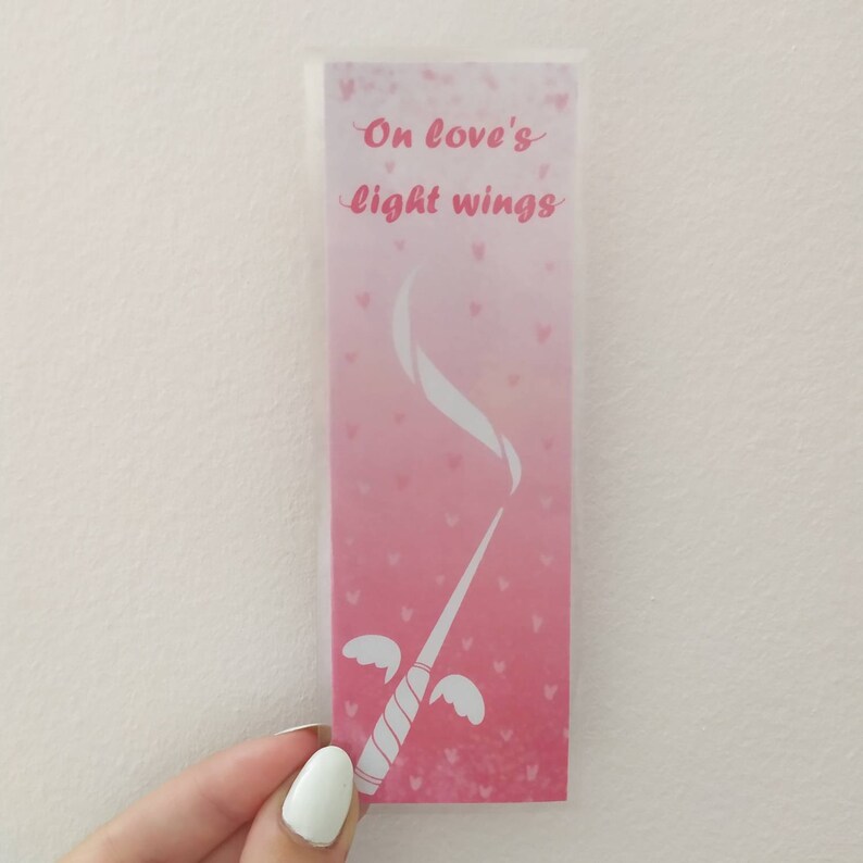 May include: Pink and white bookmark with a white wand with wings and the text "On love's light wings" in a pink heart-shaped design.