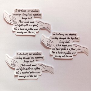 Peut inclure: Trois autocollants blancs avec du texte noir et un dessin simple de deux mains se tendant l'une vers l'autre. Le texte dit : "Dans les t&eacute;n&egrave;bres, deux ombres, atteignant &agrave; travers le d&eacute;sespoir, la lourde t&acirc;che. Leurs mains se rencontrent, et la lumi&egrave;re se r&eacute;pand en un flot, comme une centaine de bras dor&eacute;s versant le soleil."