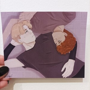 May include: Two people are lying on a patterned surface, one with blonde hair and a purple shirt, the other with red hair and a black shirt. They are facing each other and appear to be cuddling.