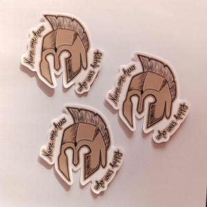 Peut inclure: Trois autocollants blancs avec une illustration brune d'un casque spartiate. Chaque autocollant porte le texte "Name one hero who was happy" &eacute;crit autour du casque.