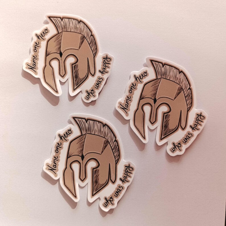 Peut inclure: Trois autocollants blancs avec une illustration brune d'un casque spartiate. Chaque autocollant porte le texte "Name one hero who was happy" &eacute;crit autour du casque.