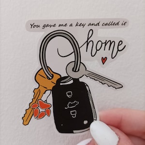 May include: A white sticker with a black and white illustration of a key chain with a car key, a house key, and a small red heart. The text "You gave me a key and called it home" is written in black at the top of the sticker.