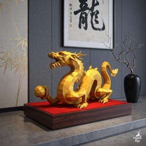 May include: A golden, low-poly geometric dragon sculpture featuring sharp, angular facets and a textured surface. The dragon is posed in a crouching position, holding a small golden sphere in one claw, and is mounted on a rectangular wooden base.