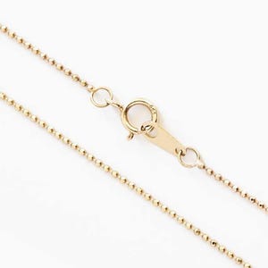 May include: A delicate gold-colored ball chain necklace with a lobster clasp. The necklace is displayed against a white background, highlighting the fine details of the jewelry. The chain is made of small, evenly spaced beads.