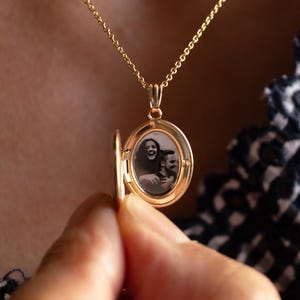 May include: A gold oval locket pendant on a delicate gold chain. The locket is open, revealing a small black and white photograph of two people inside. The metal has a polished finish, and the pendant hangs from a simple, thin gold necklace.