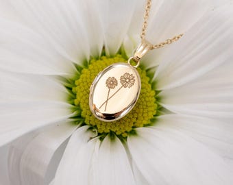May include: A gold-colored oval locket necklace with a delicate chain. The locket features an engraved floral design of two flowers. The necklace is set against a white flower with a yellow center.