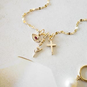 K&ouml;nnte beinhalten: Eine zarte goldene Halskette mit mehreren Anh&auml;ngern, darunter ein Auge mit rotem Stein, ein Zweig und ein Kreuz. Ein goldener Creolen-Ohrring mit einer wei&szlig;en Perle ist ebenfalls zu sehen. Der Schmuck wird auf einer wei&szlig;en Oberfl&auml;che pr&auml;sentiert.
