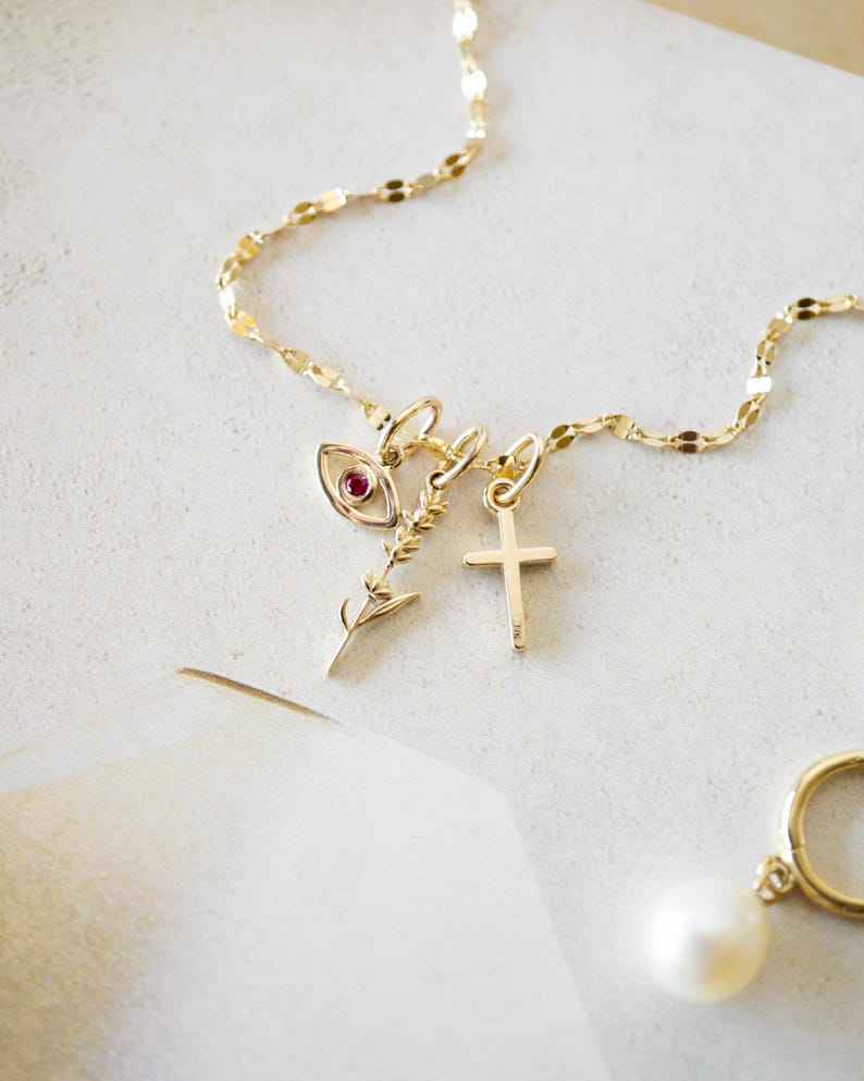 K&ouml;nnte beinhalten: Eine zarte goldene Halskette mit mehreren Anh&auml;ngern, darunter ein Auge mit rotem Stein, ein Zweig und ein Kreuz. Ein goldener Creolen-Ohrring mit einer wei&szlig;en Perle ist ebenfalls zu sehen. Der Schmuck wird auf einer wei&szlig;en Oberfl&auml;che pr&auml;sentiert.