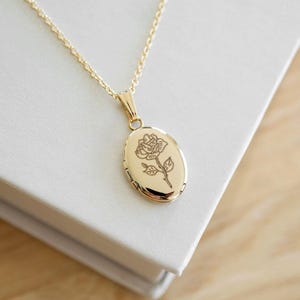 May include: A gold oval locket necklace featuring a rose engraving, suspended from a fine chain. The necklace rests on a white surface. The locket is approximately 2.5 cm in length. The background is a wooden surface.