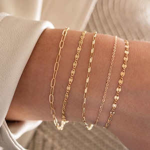 May include: Four gold chain bracelets stacked on a person's wrist. The bracelets are different styles, including a delicate chain, a chain with small oval links, and a chain with small round links.