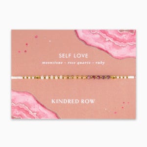 May include: A pink card with a watercolor design featuring a pink and white agate. The card says "SELF LOVE" and "moonstone - rose quartz - ruby" in white text. Below the text is a beaded bracelet with white, gold, and pink beads. The card also says "KINDRED ROW" in white text.