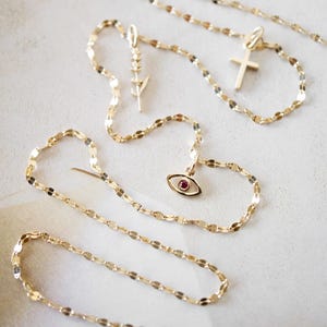 K&ouml;nnte beinhalten: Mehrere goldfarbene Halsketten mit verschiedenen Anh&auml;ngern: ein Kreuz, ein Auge mit rotem Stein und ein Blatt. Die Ketten bestehen aus kleinen, flachen Gliedern. Die Halsketten sind auf einer hellen Oberfl&auml;che angeordnet, um ihre Details hervorzuheben.