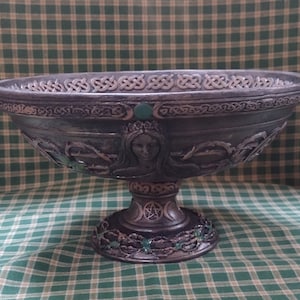 May include: A dark brown ceramic bowl with a Celtic knot design around the rim. The bowl features a raised design of a woman's face with a crown and a triple moon symbol on the base. The bowl is decorated with green gemstones.