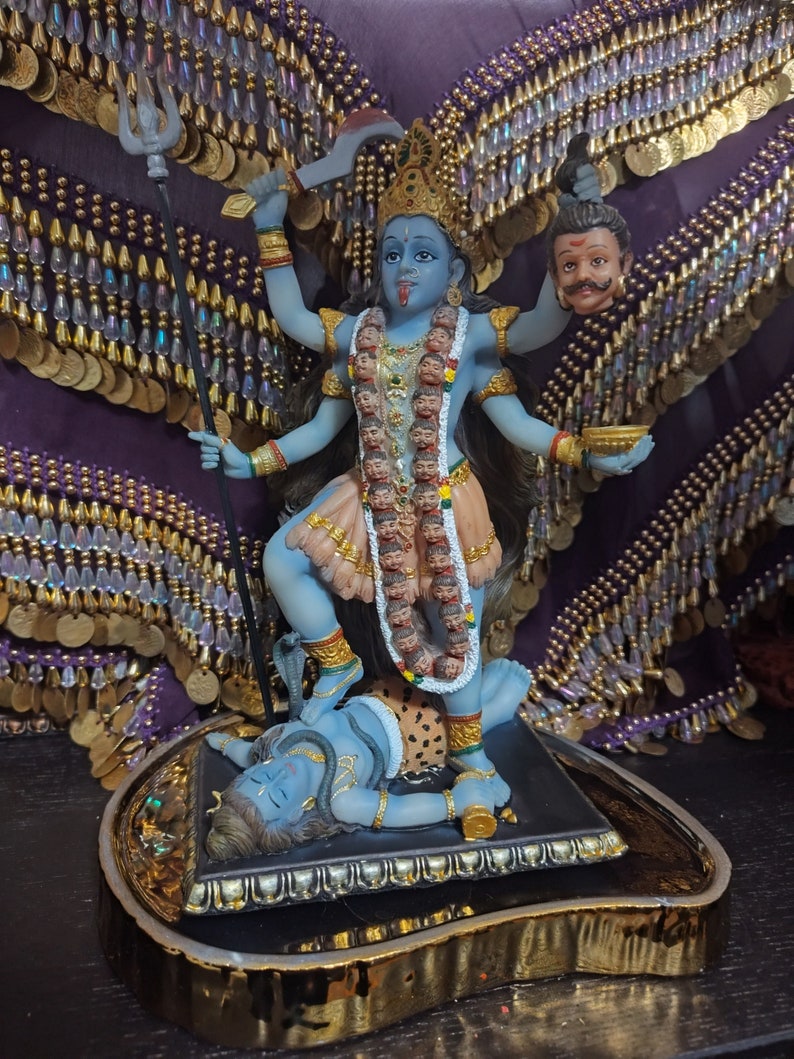 May include: A blue and peach-colored statue of a deity with multiple arms, holding various objects, and standing on a figure. The statue has a garland of heads and is adorned with gold jewelry. The base is black with gold trim.