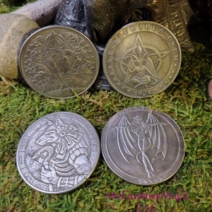 May include: Four silver-toned metal coins with different designs. The first coin features a hooded figure with a staff and a snake. The second coin features a pentagram with a figure sitting in the center. The third coin features a creature with a snake and a crown. The fourth coin features a winged figure with a tail.