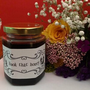 May include: A glass jar of honey with a white label that says "Hook that honey". The jar is sitting on a white surface next to a bouquet of flowers.