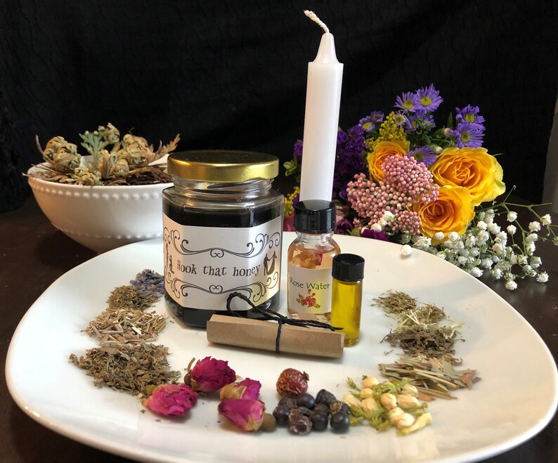 May include: A white plate containing a selection of dried herbs, flowers, and berries. On the plate are a jar of honey, a bottle of rose water, a bottle of oil, and a white candle. The label on the honey jar reads "Hook that honey".