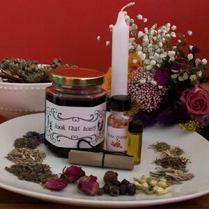 May include: A white plate containing a selection of herbs, flowers, and other ingredients for a love spell. The ingredients include dried herbs, rose petals, berries, and a small jar of honey labelled "Hook that honey". There is also a small bottle of rose water and a small bottle of oil.