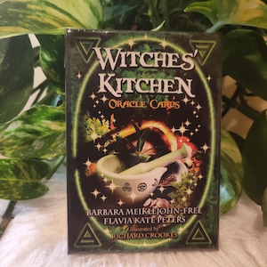 May include: A green box with a white mortar and pestle on the front. The box has a gold background with a green border and the text "Witches' Kitchen Oracle Cards" in white. The text "Barbara Meiklejohn-Free" and "Flavia Kate Peters" is in white on a black background. The text "Illustrated by Richard Crookes" is in white on a black background.