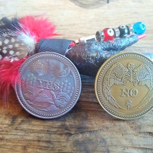 May include: Two round metal coins with opposite answers, "YES" and "NO". The "YES" coin has a silver finish and features two hands with an eye in the palm of each hand. The "NO" coin has a gold finish and features a skeleton with wings.