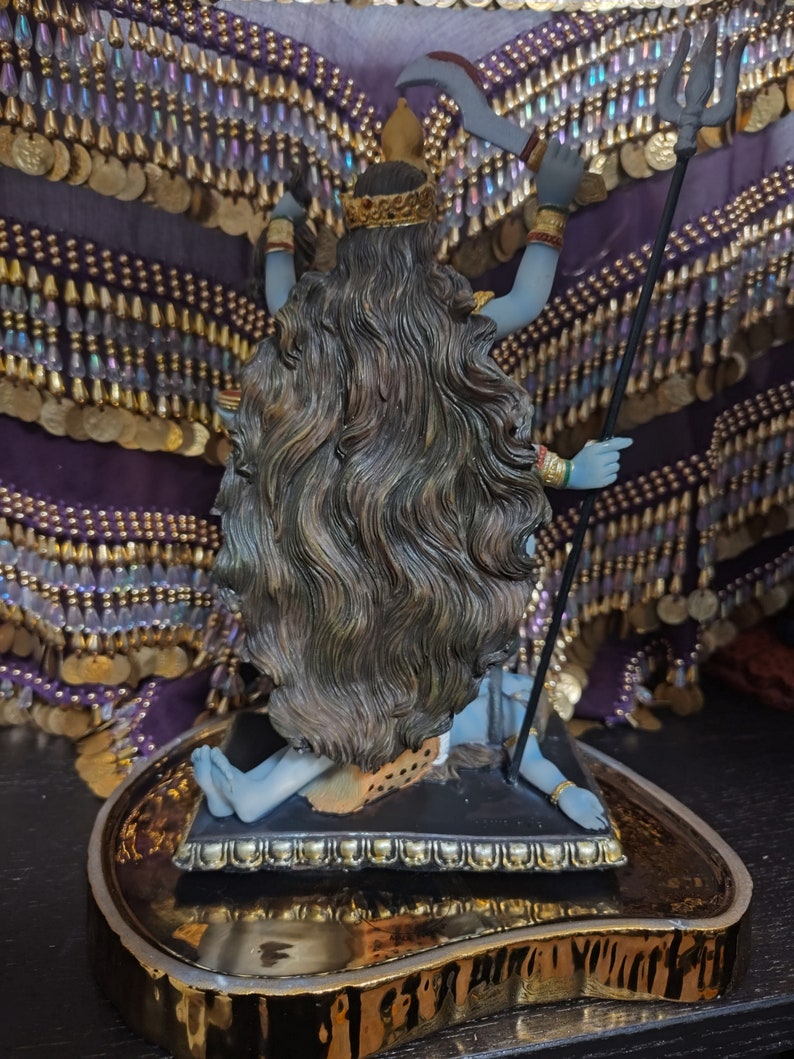 May include: A detailed statue of a deity with blue skin, long dark hair, and a golden crown. The figure holds a trident and a curved blade. The statue is on a gold-trimmed base, with a purple and gold beaded backdrop.