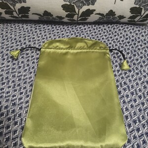 May include: A green satin drawstring pouch with black drawstrings. The pouch is lying flat on a patterned fabric surface.
