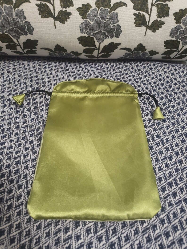 May include: A green satin drawstring pouch with black drawstrings. The pouch is lying flat on a patterned fabric surface.