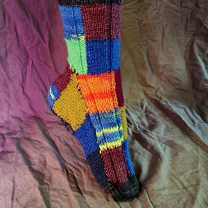 Op de afbeelding: Een gebreide sok in patchwork-stijl. De bovenste boord is donkergrijs, overgaand in een levendige mix van blauwe, groene, oranje, gele en rode vierkanten. De sok heeft een textuurpatroon.