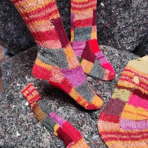 May include: Hand-knitted socks in a patchwork design. The socks feature a mix of red, orange, pink, gray, and purple hues. The image shows a pair of socks, a single sock, and a cuff, all with a striped and square pattern.