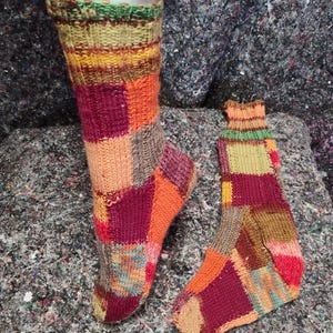 Könnte beinhalten: Ein Paar handgestrickte Socken mit Patchwork-Design. Die Socken zeigen eine Vielzahl von Farben, darunter Orange, Burgunderrot, Grün und Gelb. Die Socken werden auf einem Schaufensterbein und einem grauen Untergrund präsentiert.