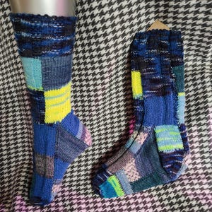 Könnte beinhalten: Ein Paar handgefertigte Socken im Patchwork-Stil. Die Socken weisen eine Vielzahl von gestrickten Quadraten in Blau, Gelb, Grün und Rosa auf. Eine Socke befindet sich auf einer Ausstellungsform, während die andere flach liegt. Der Hintergrund ist ein schwarz-weißes Hahnentrittmuster.