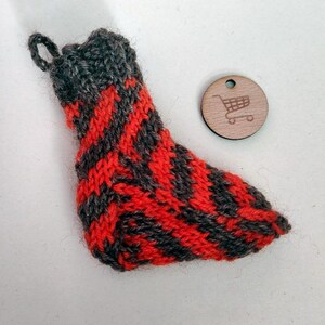 May include: A small, hand-knitted stocking in red and gray, with a loop for hanging. The stocking features a striped pattern. A small, round wooden tag with a shopping cart icon is next to the stocking.