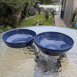 May include: Two navy blue ceramic bowls with a speckled finish. The bowls have a simple, rounded design, suitable for serving soups, salads, or other dishes. The bowls are on a glass table, with a blurred background of greenery.
