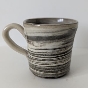 May include: A ceramic mug featuring a marbled pattern of horizontal bands in shades of charcoal grey, black, and off-white. The mug has a smooth, glossy finish, a wide circular rim, and a curved handle extending from the side of the vessel.