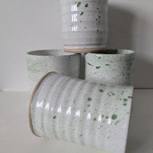 May include: A collection of four ceramic tumblers with a white glazed finish and green speckled patterns. The tumblers feature a ribbed texture along the sides and a visible tan clay base. Three tumblers are upright, while one is lying horizontally.