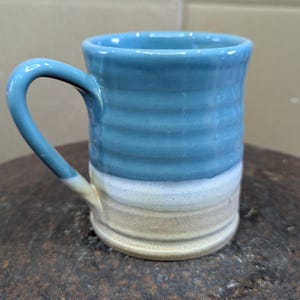 May include: A handmade ceramic mug with a blue, white, and tan colour scheme. The mug features a curved handle and horizontal textured lines around the body. The mug is approximately 10 cm tall.