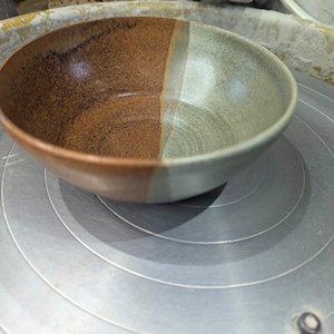 May include: A handcrafted ceramic bowl with a dual-tone aesthetic. One side is speckled brown, the other a muted green. The bowl rests on a grey surface with concentric circles, probably a potter's wheel. The interior of the bowl has a smooth, glossy finish.