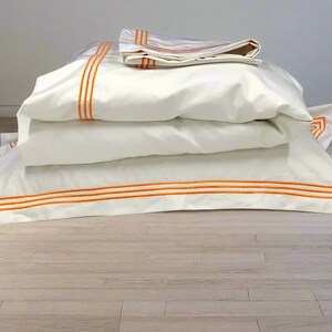 May include: A stack of white bedding with orange trim. The bedding is folded and arranged on a light wood floor.