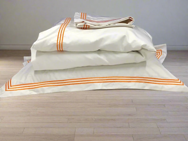 May include: A stack of white bedding with orange trim. The bedding is folded and arranged on a light wood floor.
