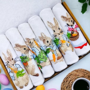 May include: Set of six white cotton tea towels with embroidered images of Peter Rabbit and his friends. Each towel features a different rabbit character holding a carrot.