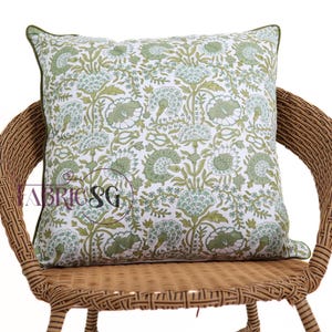 May include: A square decorative pillow with a floral pattern in shades of green and white, resting on a wicker chair. The pillow has a green border and a repeating design of stylized flowers and leaves.
