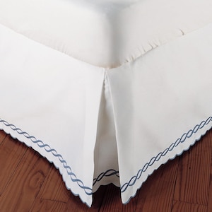 May include: White bed skirt with a blue embroidered border. The skirt has a pleated design and is shown on a wooden floor.