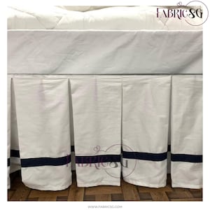 Może przedstawiać: Biała spódnica do łóżka z granatowym paskiem w pobliżu dolnej krawędzi. Spódnica ma plisowany krój i jest wykonana z gładkiej tkaniny. Górna część spódnicy do łóżka jest biała.